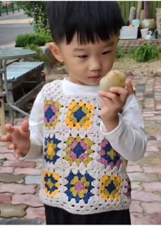 a young boy holding an apple in his hand and wearing a crocheted vest