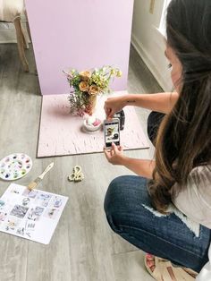 a woman sitting on the floor taking pictures with her cell phone and painting flowers in a vase