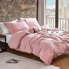 a bed with pink comforter and pillows in front of a stone wall, next to a window