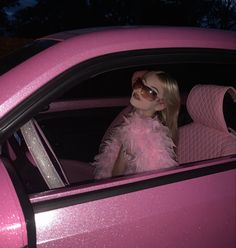 a woman sitting in the back seat of a pink car wearing sunglasses and a feathered dress