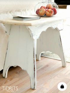 a white table with apples on top and a basket of fruit on the end, sitting in front of a couch