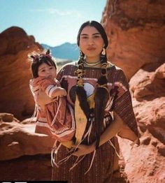 a woman holding a baby in her arms while standing next to some rocks and mountains