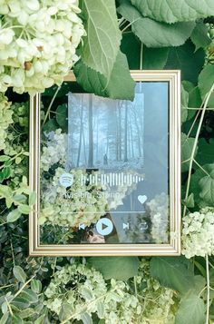 a mirror is surrounded by green leaves and white hydranges, with the reflection of a forest in it