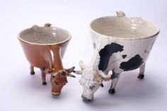 two ceramic cows standing next to each other on a white surface with one cow eating from the ground