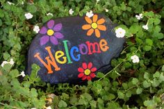 a rock with the word welcome painted on it sitting in some green grass and flowers