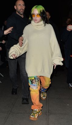 a woman with bright green hair and yellow pants is walking down the street while holding onto her skateboard