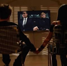 two people sitting in chairs watching tv with one person holding his hand out to the other