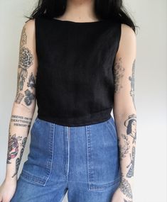 a woman with tattoos standing in front of a white wall wearing jeans and a black top