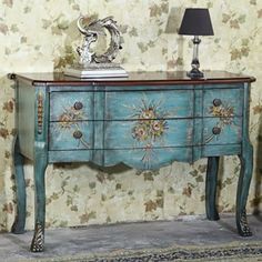 a blue painted dresser with flowers on it and a lamp next to it in front of a floral wallpapered wall