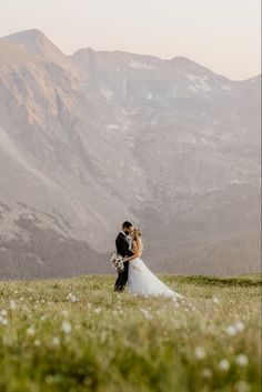 a bride and groom standing on top of a grass covered hillside with mountains in the background