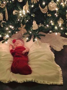 a baby in a red knitted outfit laying on a pillow under a christmas tree