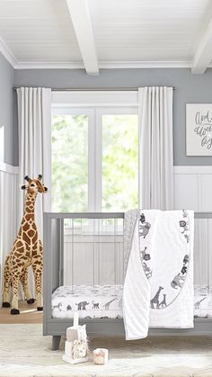 a giraffe standing next to a baby crib in a room with white curtains