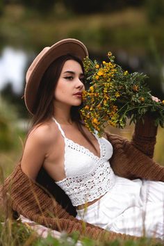 a woman sitting on the ground holding flowers in her hand and wearing a brown hat