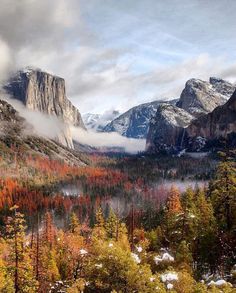 the mountains are covered in snow and surrounded by fall colored trees, with low lying clouds