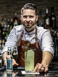 a man in an apron is behind a bar with a green drink and smiling at the camera