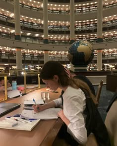 a woman sitting at a table writing in front of a globe on top of a book shelf