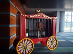 a giraffe in a red and yellow cart with wheels on the floor next to a building