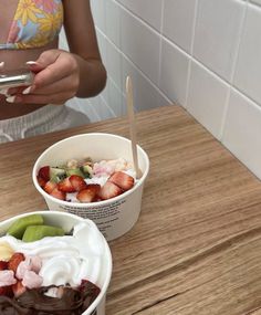 a woman holding a cell phone next to two bowls of ice cream and strawberries