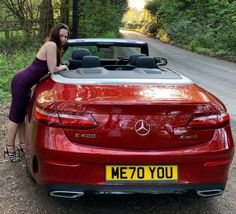 a woman leaning on the hood of a red convertible car in front of a wooded road