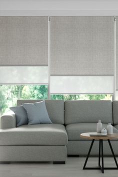 A neutral modern interior with a grey modular sofa, a round timber and black coffee table and large windows behind the sofa. The windows are covered with our double roller blinds featuring a textured grey blockout layer and a white light filtering sheer layer behind. Blinds And Curtains Together, Unique Curtains, Living Room Blinds