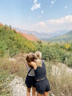 two women hugging each other while standing on a trail in the mountains with trees and grass