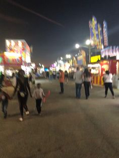 people are walking around in an amusement park at night with rides and carnival booths behind them