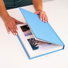 a person holding an open book on top of a white table with blue paper and photos