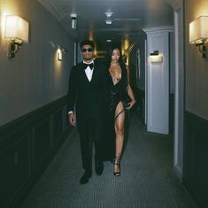 a man in a tuxedo walking down a hallway next to a woman in a black dress