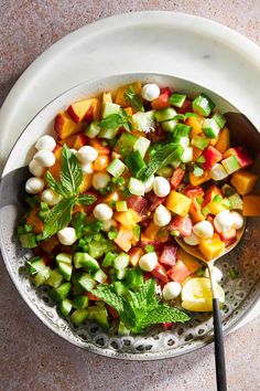 a white bowl filled with fruit and vegetables on top of a marble counter next to a metal spoon