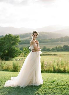 a woman in a wedding dress posing for a photo on the grass with mountains in the background