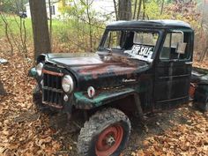 an old truck is parked in the woods with leaves on the ground next to it