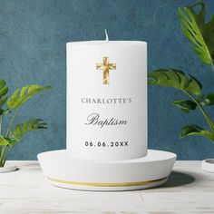 a white candle with a gold cross on it sitting next to some potted plants