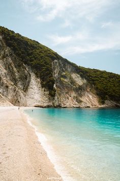 people are walking on the beach near some water and mountains in the background with blue sky