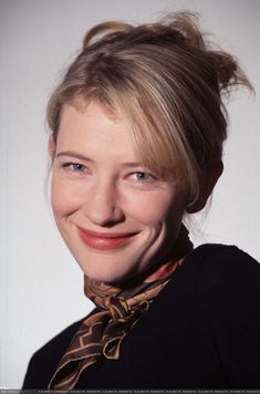 a woman with blonde hair wearing a scarf and smiling at the camera while standing in front of a white wall