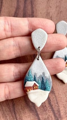 a hand holding two small white and gray earrings with houses in the woods on them