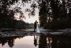 a bride and groom standing in front of a lake at sunset with trees reflected in the water