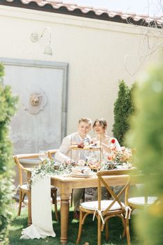 a man and woman sitting at a wooden table with flowers on it in front of a building