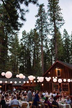 many people are sitting at tables in front of a building with paper lanterns hanging from the roof