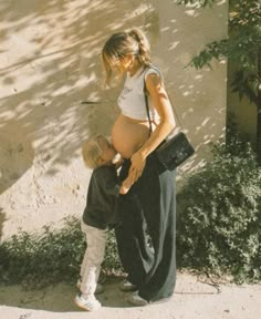 a pregnant woman holding her son's hand while standing next to a tree and building