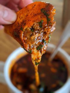 a hand holding up a piece of pizza from a bowl of chili and cheese soup