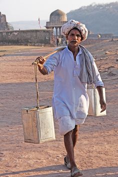 a man walking down a dirt road carrying a bucket and a stick in his hand