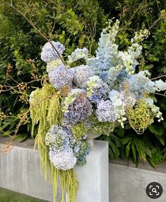 blue and white flowers are in a vase on the ground next to some shrubbery