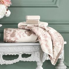 a white bench topped with lots of pillows and blankets next to a vase filled with flowers