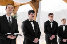 three men in tuxedos stand at the alter and look on as one man holds an open book