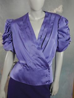 This 1980s violet purple satin blouse has a V crossover neckline edged with three pleats on each side, a fitted waistline defined with a seam, and a three-button material-covered button closure on the peplum-style waistband. The top has elegant gathered short puffed sleeves. Bust = 40 inches (101.6cm) Shoulders = 16.5 inches (41.91cm) Waist = 33 inches (83.82cm) Hips = 42 inches (106.68cm) Sleeve Length = 12 inches (30.48cm) Top Length = 23.5 inches (59.69cm) Size: 12 US, 16 UK Material: Polyest Purple Collared Blouse With Button Closure, Purple Satin Blouse, Purple V-neck Formal Blouse, Violet Blouse, 1980s Fashion Purple, Purple Button-up Blouse For Formal Occasions, Vintage Purple Tops With Button Closure, Peplum Styles, Purple Blouse