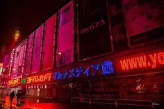 people are walking down the street in front of a building with neon lights on it