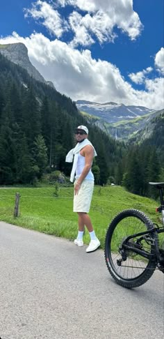 a man standing next to a bike on the side of a road in front of mountains