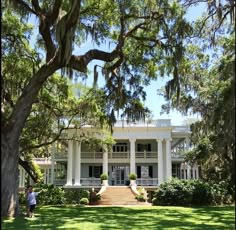 a large white house surrounded by trees and green grass with two people standing in front of it