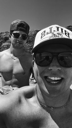 two shirtless men sitting on the beach with one wearing a baseball cap and sunglasses