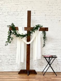 a cross with greenery draped over it in front of a white brick wall and small table